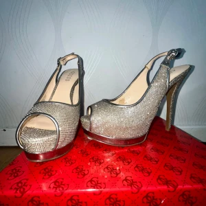 Glittriga silverfärgade pumps från Guess - Säljer ett par snygga pumps från Guess i glittrigt silver med öppen tå och hög klack. Skorna har en elegant design med metalliska detaljer och plattformssula. Perfekta för att sticka ut och ge outfiten extra glamour.