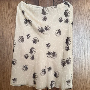 Beige kjol med svart mönster - Säljer en beige kjol med svart blommigt mönster och lätt genomskinligt yttertyg. Kjolen har en rak passform. Perfekt för dig som gillar en stilren men ändå unik look.