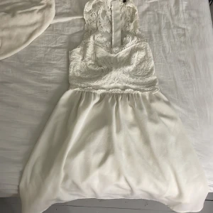 Vit spetsklänning med ärmlös design - Säljer en elegant vit klänning med spets upptill och ärmlös design. Klänningen har en vacker v-ringning och öppen rygg, samt en lätt utställd kjol i mjukt tyg. Perfekt för sommarens speciella tillfällen.