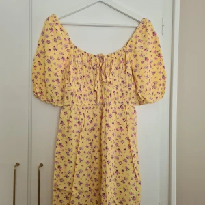 Gul blommig klänning med puffärm från Bikbok - Söt gul klänning med lila och rosa blommor, puffärmar och smockad överdel. Knytning framtill och lätt rynkat tyg ger en romantisk känsla. 