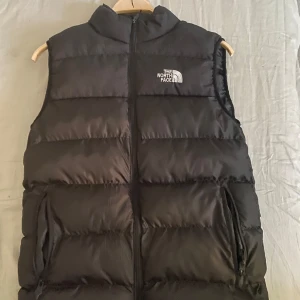 Svart dunväst från The North Face - Säljer en svart dunväst från The North Face med dragkedja framtill och två sidofickor med dragkedja. Västen har en klassisk puffer-design och logga på bröstet. Perfekt för lager-på-lager under kyliga dagar.
