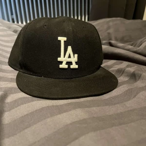 Svart LA-keps från New Era 59FIFTY - Säljer en svart keps från New Era 59FIFTY med LA-logga framtill i vitt. Kepsen har klassisk rak skärm och broderade detaljer, inklusive MLB-logga bak och New Era-logga på sidan. Perfekt för dig som gillar sportig stil. Knappt använd 