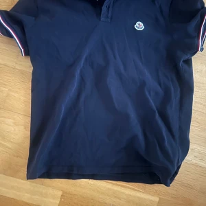 Marinblå pikétröja från Moncler - Säljer en marinblå pikétröja från Moncler med korta ärmar och klassisk krage. Tröjan har Moncler-logga på bröstet och rödvita detaljer på ärmsluten. Perfekt för en stilren look.