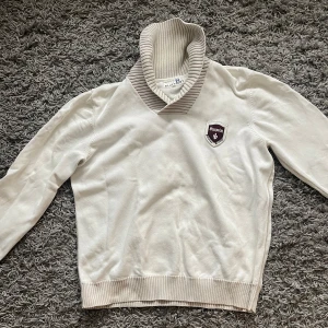 Vit polotröja från Blueprint - Säljer en stilren vit polotröja från Blueprint med ribbad beige krage och muddar. Tröjan har ett broderat märke på bröstet och är långärmad. Perfekt för dig som gillar klassisk och finare stil