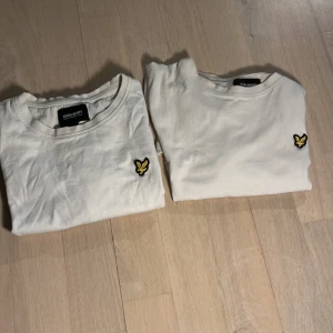 Vit t-shirt från Lyle & Scott Junior - Säljer två klassiska vita t-shirts från Lyle & Scott Junior med den ikoniska gula loggan broderad på bröstet. T-shirtsen har rund hals och korta ärmar. Perfekt för en stilren och enkel look.