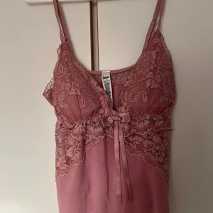 Rosa spetslinne med rosett - Säljer ett romantiskt rosa linne med spetsdetaljer och en söt rosett framtill. Linnet har smala axelband och v-ringning, samt vacker spets både fram och bak. Perfekt för en feminin look.