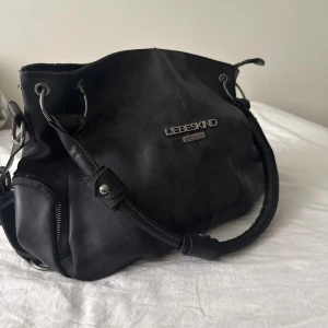 Svart handväska från Liebeskind - Säljer en svart handväska från Liebeskind med silverfärgade metalldetaljer och dragkedja på sidan. Väskan har rundade former och breda handtag, perfekt för dig som vill ha en stilren och rymlig accessoar.