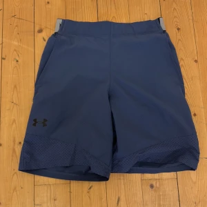 Blå träningsshorts från Under Armour - Mörkblå träningsshorts från Under Armour med elastisk midja och stor logotyp på benet. Shortsen har ventilerande meshdetaljer nedtill och snörning i midjan för extra komfort. Perfekta för gym eller löpning.