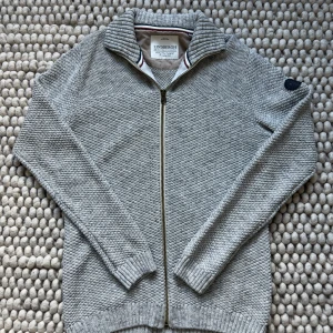 Grå stickad kofta från Lindbergh - Säljer en grå stickad kofta från Lindbergh med dragkedja framtill och hög krage. Tröjan har lång ärm och ribbade muddar vid ärmslut och nederkant. Perfekt för lager-på-lager och stilren vardagslook.