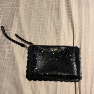 Svart kuvertväska från Victoria's Secret - Säljer en svart kuvertväska från Victoria's Secret med snygga utskurna detaljer och vågig kant. Väskan har dragkedja och handledsrem. Helt ny!