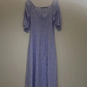 Ljuslila långklänning med blommigt mönster - Superfin ljuslila långklänning med vitt blommigt mönster. Klänningen har puffiga trekvartsärmar, v-ringning och en slits framtill. Perfekt för sommarens alla tillfällen! Materialet känns lätt och luftigt.