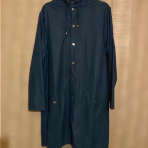 Blå regnjacka från Rains - Säljer en stilren blå regnjacka från Rains med långa ärmar och knappar framtill. Jackan har en huva med snörning och praktiska fickor. Perfekt för regniga dagar!