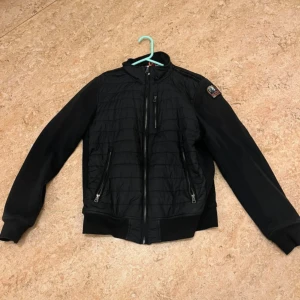 Svart jacka från Parajumpers - Säljer en svart jacka från Parajumpers med quiltad design och dragkedja framtill. Jackan har långa ärmar och en broderad logotyp på ärmen. Perfekt för kyligare dagar.