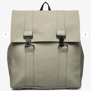 Beige ryggsäck från Rains - Snygg och stilren beige ryggsäck från Rains med två svarta spännen framtill. Tillverkad i ett syntetmaterial som ger en modern look. Ryggsäcken har justerbara axelremmar och ett rymligt huvudfack. Perfekt för både vardag och utflykter.