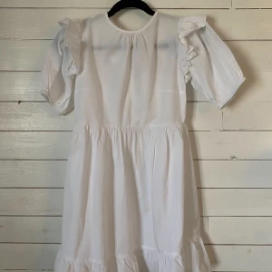 Vit klänning med volanger - Vit klänning med volanger och korta ärmar. Den har en öppen rygg med knytband och volangdetaljer. Perfekt för student!