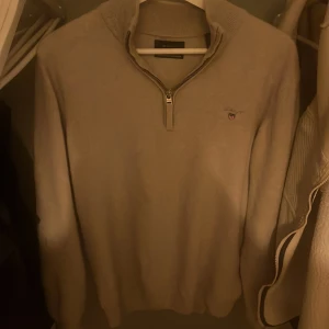 Grå ulltröja från GANT - Säljer en stilren beige tröja från GANT i superfin lammull. Tröjan har en dragkedja vid halsen och ett diskret broderat emblem på bröstet. Perfekt för kyliga dagar!