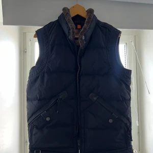 Svart dunväst från Parajumpers - Säljer en snygg svart dunväst från Parajumpers Masterpiece Series. Västen har en mysig krage med pälsdetaljer och praktiska fickor med knappar. Perfekt för kyliga dagar! Ligger på ett värde runt 5000kr idag