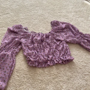 Lila blommig blus - Säljer en söt lila blommig blus med långa ärmar och volangdetaljer. Blusen har en elastisk midja och är perfekt för en romantisk look. Den är gjord i ett lätt och luftigt material.