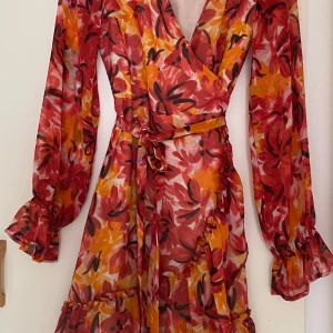 Blommig klänning från Gina Tricot - Säljer en färgglad blommig klänning från Gina Tricot. Klänningen har långa ärmar och ett omlottmönster med volangdetaljer. Perfekt för sommarens fester!