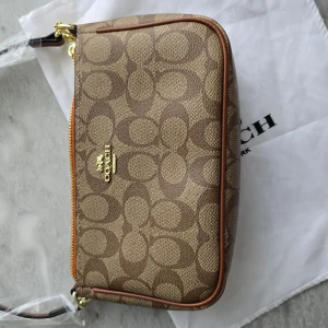 Beige och brun axelväska från Coach - Säljer en snygg axelväska från Coach i beige och brunt med deras ikoniska mönster. Väskan har en dragkedja upptill och guldfärgade detaljer. Perfekt för att bära dina viktigaste saker med stil.