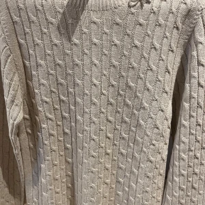 Beige stickad tröja med rosett - Säljer en stilren beige stickad tröja med ett snyggt flätmönster och en glittrig rosett vid halsen. M/L ordinarie pris ca1800kr helt ny