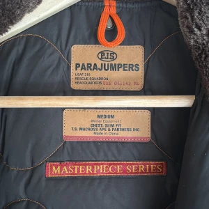 Svart vinterjacka från Parajumpers - Säljer en svart vinterjacka från Parajumpers med pälsdetaljer på huvan och en cool patch på ärmen. Jackan har en dragkedja och knappar framtill samt flera praktiska fickor. Perfekt för kalla dagar!