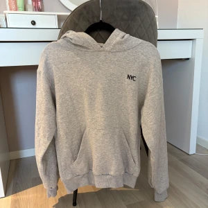Grå hoodie  - Säljer en stilren grå hoodie metryckt på bröstet. Den har en klassisk känguruficka och en bekväm huva. Perfekt för en avslappnad look.