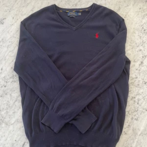 Mörkblå tröja från Ralph Lauren - Säljer en stilren mörkblå tröja från Ralph Lauren med v-ringning och det klassiska röda logotypbroderiet på bröstet. Tröjan har långa ärmar och är tillverkad i bomull för en bekväm passform. Perfekt för en avslappnad men ändå elegant look.