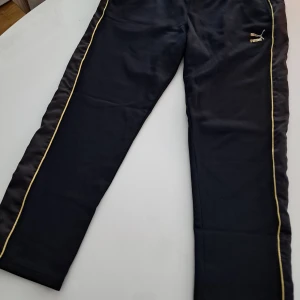 Svarta träningsbyxor från Puma * Kenza - Snygga svarta träningsbyxor från Puma med gulddetaljer längs sidorna. Byxorna har en dragkedja på baksidan och elastisk midja för extra komfort. Perfekta för en aktiv livsstil eller en avslappnad dag.
