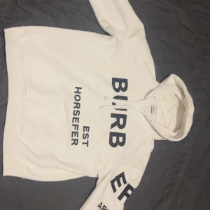Burberry Hoodie - Säljer en vit hoodie från Burberry med svart texttryck på framsidan och ärmarna. Hoodien har en klassisk känguruficka och justerbar dragsko i huvan. Perfekt för en stilren och avslappnad look.
