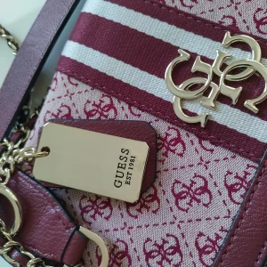 Axelväska från Guess - Snygg axelväska från Guess i vinrött och vitt med monogrammönster. Väskan har en kedjedetalj på axelremmen och ett stort metallogo framtill. Perfekt för att ge din outfit en lyxig touch.