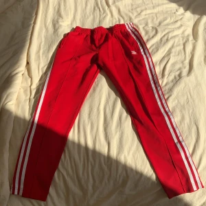 Röda träningsbyxor med vita ränder - Snygga röda träningsbyxor från Adidas med klassiska vita ränder längs sidorna. Byxorna har en elastisk midja för extra komfort och en liten logga på ena benet. Perfekta för sport och fritid.