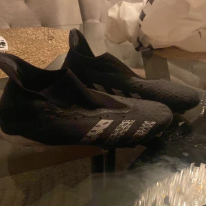 Svarta fotbollsskor Adidas Predator - Säljer ett par svarta Adidas Predator fotbollsskor med vita detaljer. Skorna har en strömlinjeformad design med dobbar för optimalt grepp på planen. Perfekta för fotbollsspelare som vill ha både stil och funktion.