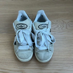 Beige Adidas Campus sneakers - Säljer ett par snygga beige Adidas Campus sneakers i mocka med svarta detaljer och vita snören. Använda en sommar!