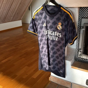 Svart Real Madrid fotbollströja från Adidas - Snygg svart Real Madrid fotbollströja från Adidas med ett vågigt mönster och gula detaljer. Tröjan har korta ärmar och klubbens emblem på bröstet. Perfekt för alla fans som vill visa sitt stöd med stil. Jag köpte den för 1100 men är för för liten för mig nu så säljer den för ett billigt pris!