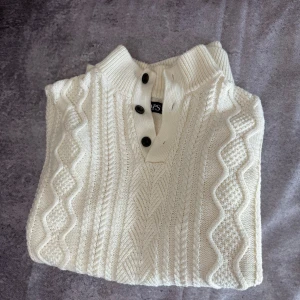 Vit stickad tröja från Chaps - Säljer en stilren vit stickad tröja från Chaps med ett klassiskt flätmönster. Tröjan har långa ärmar och tre knappar vid halsen för en snygg detalj. Perfekt för kyligare dagar.