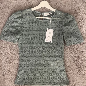 Grön spetsblus från VILA - Säljer en elegant grön spetsblus från VILA med korta puffärmar och rund hals. Blusen har ett vackert spetsmönster och en knappdetalj i nacken. Perfekt för att ge en romantisk touch till din outfit.