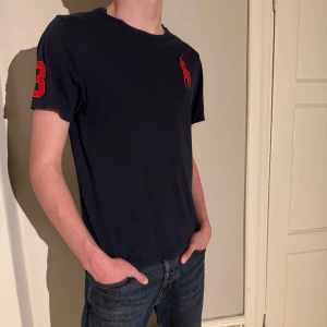 Mörkblå t-shirt från Polo Ralph Lauren - Snygg mörkblå t-shirt från Polo Ralph Lauren med röd broderad logga på bröstet och siffran 3 på ärmen. Perfekt till våren och sommaren. Modellen på bilden är 176cm.