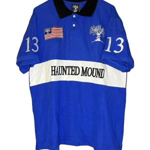 Blå pikétröja från Haunted Mound - Säljer en blå pikétröja från Haunted Mound med svart krage och vita detaljer. Tröjan har nummer 13 på ärmarna och ett tryck av en flagga och ett träd på bröstet. Texten 'Haunted Mound' är tryckt över bröstet. Perfekt för en avslappnad stil.