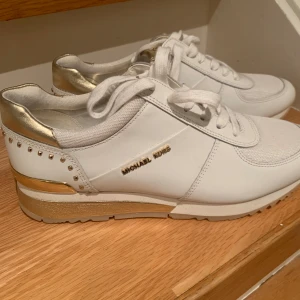 Vita sneakers från Michael Kors stl 38 - Snygga vita sneakers från Michael Kors med gulddetaljer och snörning. Skorna har en elegant design med guldfärgade inslag på hälen och logotypen på sidan. Perfekta för en stilren look. Skicket är väldigt bra, aldrig använda! 