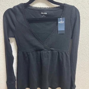 Svart omlottopp från Hollister - Säljer en stilren svart omlottopp/babydoll topp från Hollister med v-ringning och spetsdetaljer.           Toppen har långa ärmar och en bekväm passform som passar perfekt till både jeans och kjol.                                                             Man kan ha ett linne under tröjan så de sticker ut lite så blir det också superfint!                                           Perfekt för en chic och avslappnad look.  Stor i storlek, mer som i storlek S! 