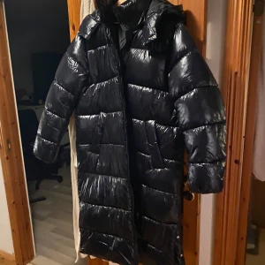 Svart lång dunjacka med huva - Säljer en svart lång dunjacka med huva. Jackan har en glansig yta och är perfekt för kalla dagar. Jackan är helt oanvänd. 