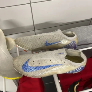 Vita fotbollsskor från Nike - Snygga vita fotbollsskor från Nike med en glittrig blå swoosh. Skorna har en modern design utan snörning och är perfekta för fotbollsplanen. De har en strömlinjeformad form och är gjorda för att ge bra grepp och kontroll.