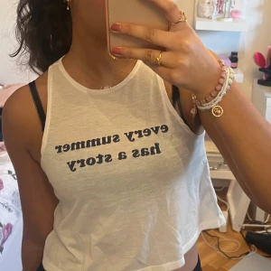 Vit ärmlös linnetopp med text - Säljer en vit ärmlös linnetopp från Divided med texten 'every summer has a story' i blått på framsidan. Perfekt för varma sommardagar. Toppen har en lös passform och är tillverkad i ett lätt och bekvämt material.