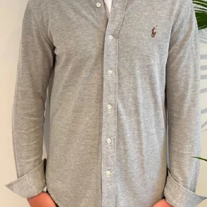 Grå långärmad skjorta från Ralph Lauren - Snygg grå långärmad skjorta från Ralph Lauren med klassisk krage och knappar framtill. Skjortan har en broderad logotyp på bröstet och är perfekt för en stilren look.