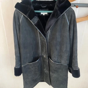 Svart mockajacka med huva - Snygg svart mockajacka med mjuk pälsfodrad huva och knappar framtill. Jackan har långa ärmar och stora fickor, perfekt för kyliga dagar. En stilren och tidlös design som passar till många tillfällen.