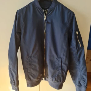 Blå bomberjacka från H&M med dragkedja - Snygg blå bomberjacka från H&M med dragkedja framtill och en ficka med dragkedja på ärmen. Jackan har ribbade muddar vid ärmslut och nederkant för en bekväm passform.  Storlek 170. Kan hämtas i Vallentuna.