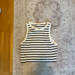 Randig ärmlös topp - Säljer en stilren ärmlös topp med svarta och vita ränder. Perfekt för varma dagar och enkel att matcha med olika outfits. Toppen har en klassisk rund halsringning. Croppad.