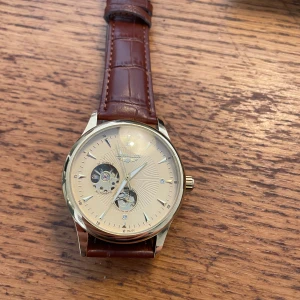 Elegant klocka med brun läderrem automatisk klocka - Säljer en elegant klocka med brun läderrem och guldtonad boett. Urtavlan är ljus med synliga mekaniska delar och blå detaljer. Baksidan är transparent och visar urverket. Perfekt för den som gillar stilrena och klassiska accessoarer.