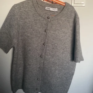 Grå kofta från Zara - Säljer en stilren grå kofta från Zara med korta ärmar och knappar framtill. Perfekt för lager-på-lager under svalare dagar. Enkel och elegant design som passar till många olika outfits.